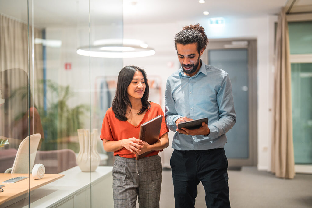 Two professionals walking through a modern office, discussing employee matters and reviewing information on a digital tablet and notebook.