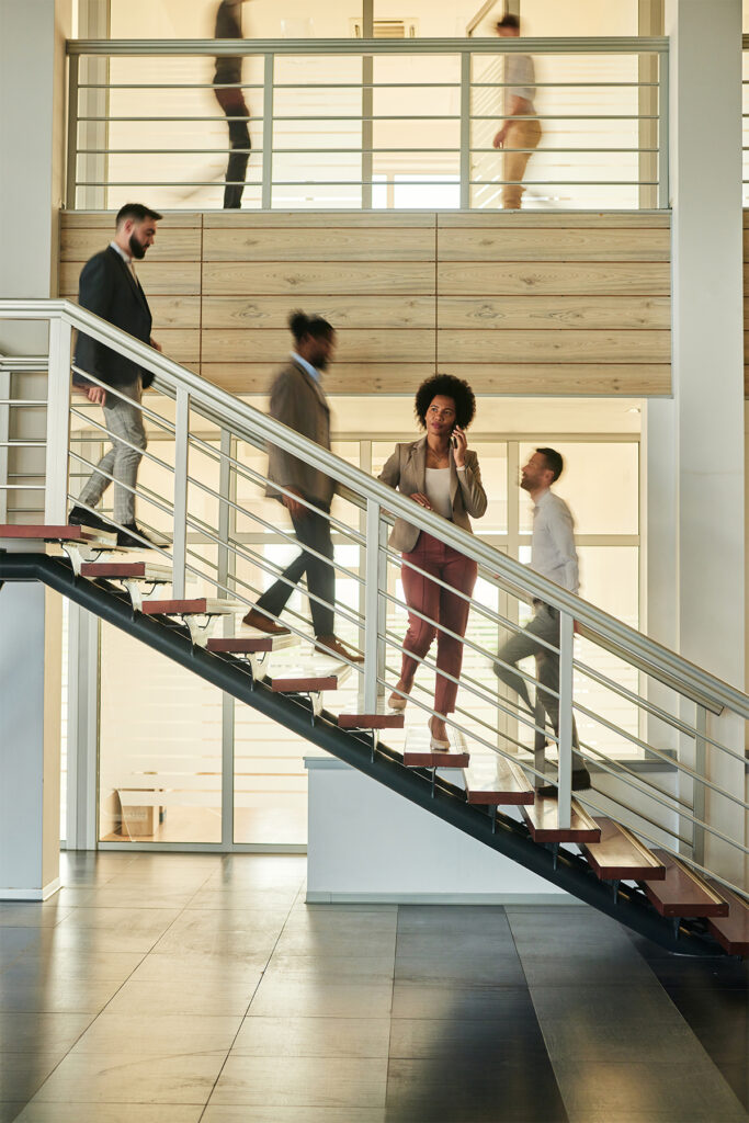 Businesswoman talking on mobile phone while standing on staircase in a hallway of an office building while her colleagues are in blurred motion.